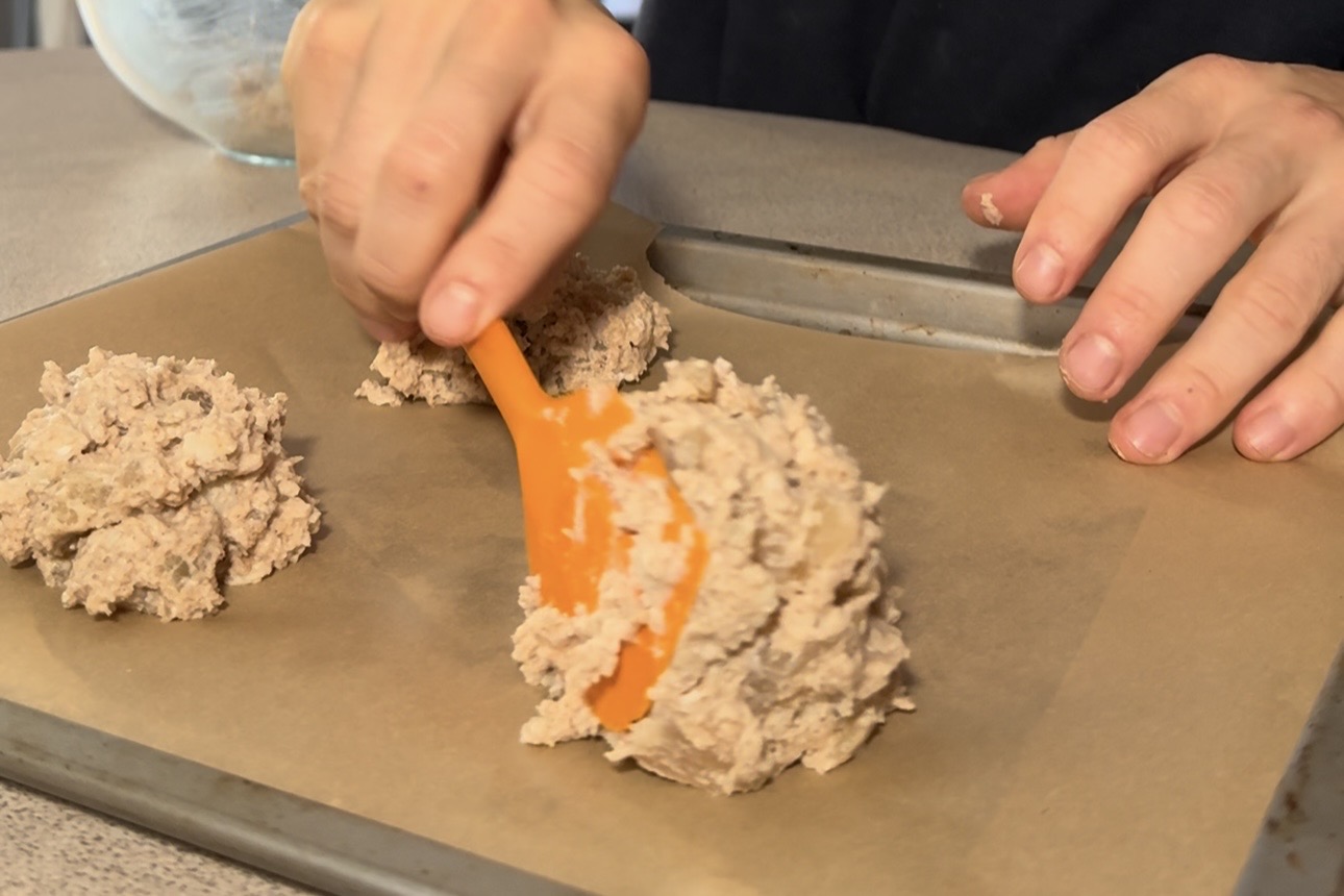 Four apple fritter batter mounds spaced apart on a parchment-lined tray